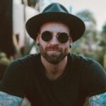 Stylish man with beard and tattoos wearing sunglasses and a hat, enjoying outdoors.
